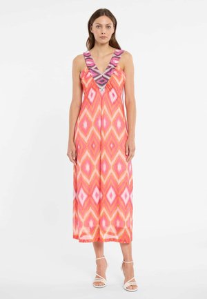 Femme portant une robe midi sans manches à col en V avec un motif losange rose, orange et blanc, associée à des talons blancs à lanières, debout sur fond blanc.
