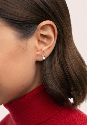 Pendientes de media luna plateada con una pequeña piedra preciosa clara, llevados en la oreja de una modelo, con un diseño elegante sobre un jersey de cuello alto rojo.