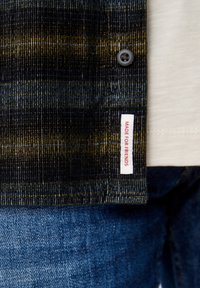 Camisa de tela a rayas negra y dorada con una superficie texturizada, que presenta un cierre de botones y una etiqueta que dice "HECHO PARA AMIGOS". Fondo de denim.