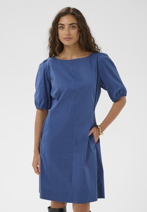 Käärus juustega naine, kes kannab sinist põlvpikkust kleiti täis varrukatega, seisab ühe käega taskus ühevärvilise tausta ees.