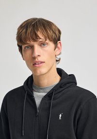 Svart zip-up huvtröja med dragsko, med en liten vit logotyp på bröstet och bärs över en grå t-shirt med rund hals. Slät tygstruktur.