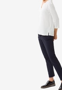 Blouse blanche à manches longues avec un col en V et des fentes sur les côt és, associée à un pantalon tailleur navy et des chaussures à lacets noires et brillantes.