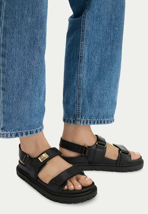 Fødder iført sorte lædersandaler med justerbare remme kombineret med blå denimjeans mod en lys baggrund.