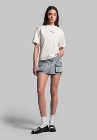 T-shirt bianco in cotone con maniche corte e logo, abbinato a una mini gonna in denim blu chiaro con due tasche anteriori. Scarpe nere e calzini bianchi.