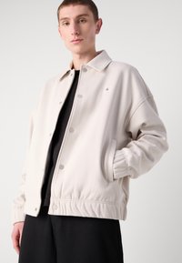 Giovane che indossa una giacca button-up beige chiaro sopra un top nero, con una mano in tasca, in posa su uno sfondo semplice.