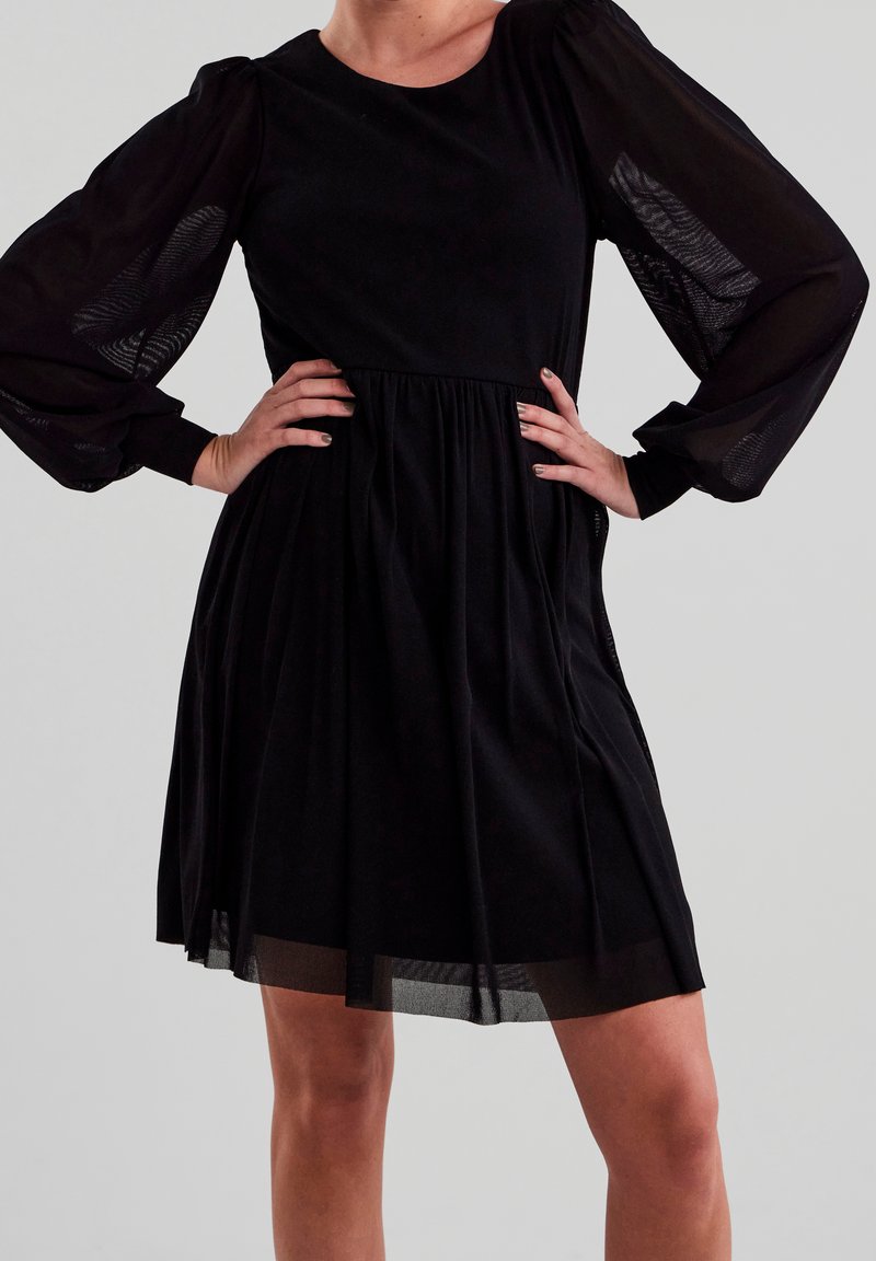 Femme portant une courte robe noire avec des manches longues bouffantes en tulle transparent, mains sur les hanches, debout devant un fond gris clair uni.