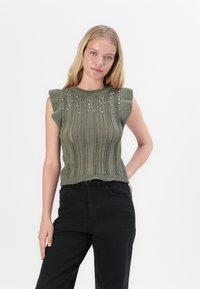 Top verde tricotat fără mâneci, cu nervuri verticale, ornamentat cu mărgele decorative și dintr-un material moale și texturat. Asortat cu blugi negri.