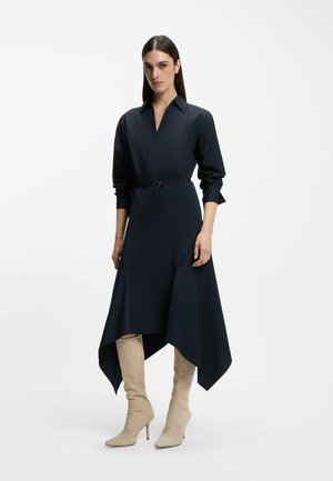 Mujer vestida con un vestido negro de manga larga con dobladillo asimétrico y botas beige de tacón hasta la rodilla, posando frente a un fondo blanco.