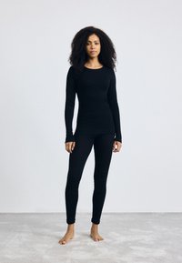 Zwart langemouwen top en strakke leggings, gemaakt van soepele stof, met een eenvoudig ontwerp en een zachte textuur. Blootsvoets op een licht tapijt.