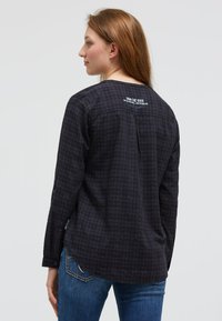 Frau mit heller Haut und langen braunen Haaren trägt ein langärmeliges Hemd mit dunklem Karomuster und weißem Text auf dem oberen Rücken sowie eine Jeans, sie ist nach links gerichtet.