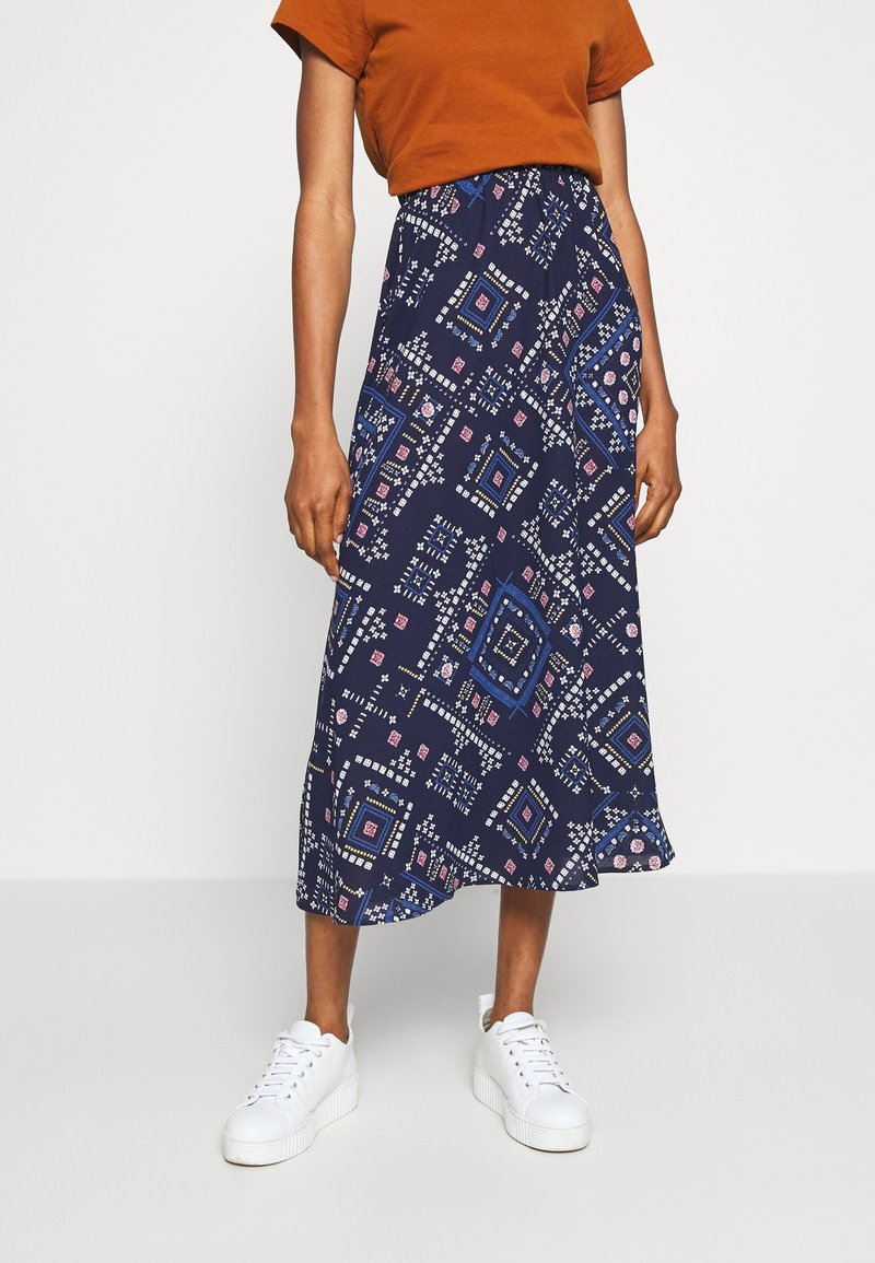 Personne portant une jupe midi bleu marine avec des motifs géométriques blancs et roses, des baskets blanches et un top orange à manches courtes, debout sur un fond uni.