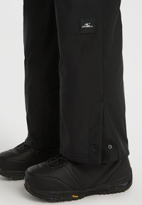Zwarte sneeuwbroeken met een gladde textuur, voorzien van een logopatch en drukknoopsluitingen bij de manchetten, gecombineerd met zwarte waterdichte boots.