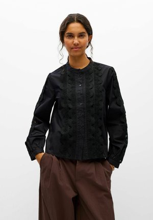 Une femme porte un chemisier noir boutonné avec des empiècements en dentelle et un pantalon marron sur un fond blanc uni.