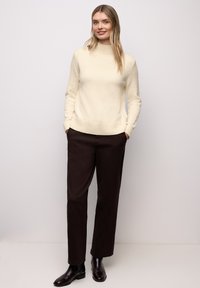 Cremefarbener Strickpullover mit hohem Ausschnitt, kombiniert mit dunkelbraunen Hosen und glänzenden schwarzen Stiefeletten. Strukturierter Stoff, lockere Passform.