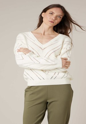 Vrouw met lang bruin haar, gekleed in een wit, opengewerkt V-hals truitje en olijfgroene broek, staand met gekruiste armen tegen een neutrale achtergrond.