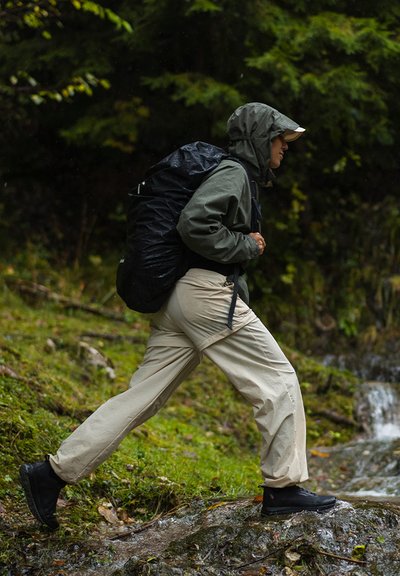 Outdoor gear featuring a black waterproof backpack, olive green jacket, beige cargo pants, and black hiking boots. Wet landscape background.
