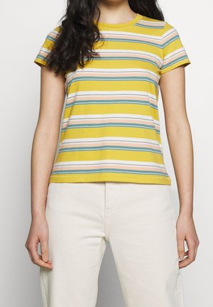 Mujer con camiseta amarilla de manga corta con rayas horizontales en tonos pastel combinada con pantalones color marfil, de pie frente a un fondo liso.