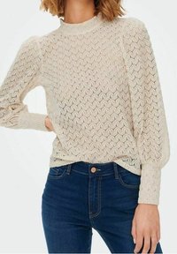 Top a maniche lunghe color beige con un motivo in pizzo, collo alto e maniche a sbuffo, abbinato a jeans blu scuro con chiusura a bottone.