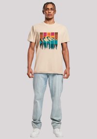 Beige katoenen t-shirt met een grafisch ontwerp van bergen en bomen in blauw, oranje en roze. Gecombineerd met lichtblauwe jeans en witte sneakers.