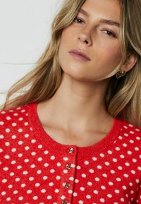 Punainen neuletakki, jossa on valkoisia pilkkuja, pyöreä pääntie ja kultaiset nappikoristeet. Materiaali vaikuttaa pehmeältä ja teksturoidulta.