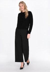 Zwarte cardigan van zacht materiaal met een ronde hals, frontknopen en een aansluitende pasvorm. Gepaard met zwarte wijde broek en hakken.