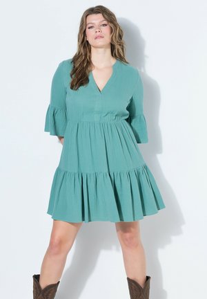 Kvinde med bølget hår iført en teal, lagdelt knælange kjole og brune cowboystøvler, står med benene fra hinanden mod en hvid baggrund.
