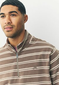 Brun og beige stribet zip-up skjorte med høj krave, der har en sølvlynlås og broderet logo på venstre bryst. Blød tekstur.