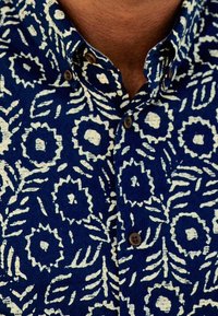 Košile s modrým květinovým vzorem a bílými motivy, s límečkem na knoflíky a hnědými knoflíky. Materiál vypadá texturovaně a má mírně tkaný vzhled.