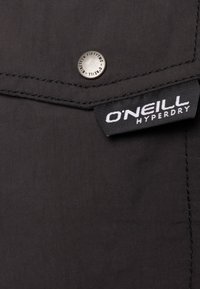 Tessuto nero con una superficie liscia, caratterizzato da un bottone a pressione argento e un'etichetta nera con testo bianco che recita "O'Neill Hyperdry."