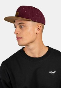 Gorra marrón con puntos blancos, visera marrón y un parche de cuero. Sudadera negra con cuello de canalé. Perfil lateral de un modelo masculino.