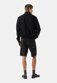 Giacca bomber nera con colletto alto, abbinata a pantaloni corti in denim neri. I pantaloni corti presentano una toppa con logo triangolare sulla tasca posteriore.