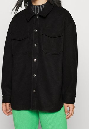 Vrouw draagt een zwart jasje met knopen en twee borstzakken over een zwart-grijs gestreept shirt en felgroene broek.