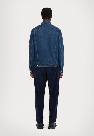 Denim jacket in dark blue with red floral embroidery on sleeves, featuring snap buttons and a tailored fit, paired with navy trousers and black shoes.