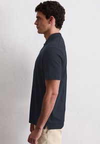 Dunkelgraues Poloshirt aus weichem Material, mit kurzen Ärmeln und einem Kragen mit drei Knöpfen. Verfügt über seitliche Schlitze am Saum für eine angenehme Bewegungsfreiheit.