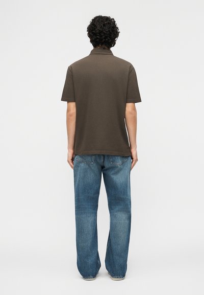 Homme aux cheveux bouclés portant un polo à manches courtes de couleur olive foncé et un jean bleu ample, debout de dos sur un fond uni.