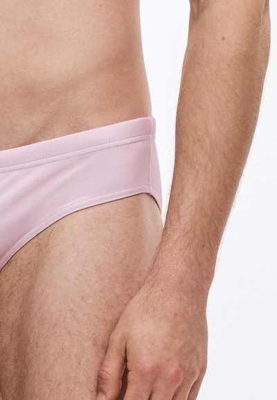 Heledast roosad ujumispüksid, millel on sile tekstuur, lai elastne vöökoht ja kahepoolse õmblusega saaled. Näha on mehe ülakeha ja käsi.