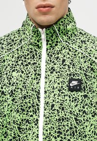 Κοντινό πλάνο ενός ατόμου που φοράει πράσινο και μαύρο μπουφάν Nike Air με λευκό φερμουάρ και λογότυπο στο στήθος, στέκεται μπροστά σε ανοιχτό φόντο.