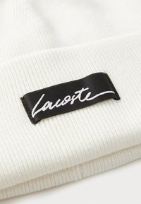 Țesătură albă cu fante, pliată, cu un etichetă dreptunghiulară neagră cusută, având text cursiv alb "Lacoste" pe etichetă.