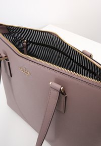 Vaaleanlila olkalaukku, jossa on teksturoitu pintakäsittely, kultainen vetoketju ja musta-valkoinen raidoitusvuori. Laukussa on myös sisätaskut järjestelyn helpottamiseksi.