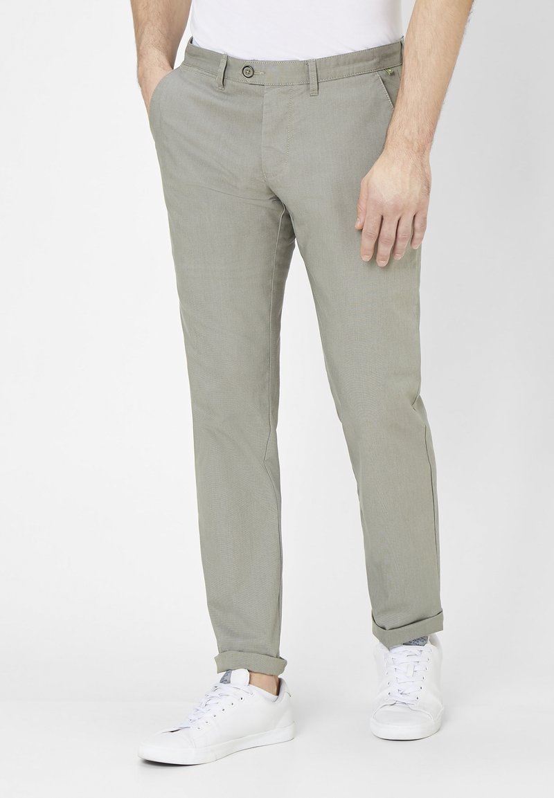 Homme portant un pantalon slim gris clair et des baskets blanches, debout les mains dans les poches sur fond blanc.