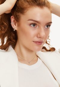 Silberner Schmuck mit Herz- und Schmetterlingsmotiven; das Modell trägt geschichtete Halsketten über einem weißen Rippoberteil mit einem weißen Blazer.