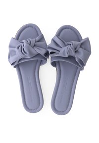 Een paar lavendel flat slide sandalen met grote geknoopte strikken op de banden en een gevoerde voetbed, gezien van bovenaf op een witte achtergrond.