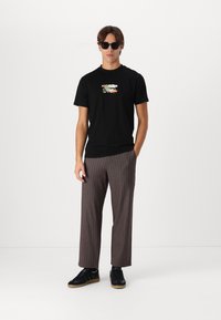 Tricou negru cu un grafic colorat pe piept, asociat cu pantaloni cu dungi maronii și teniși negri. Modelul poartă ochelari de soare întunecați.