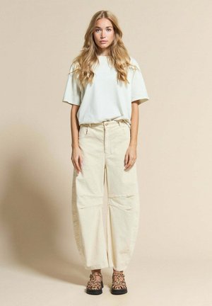 Jeune femme aux longs cheveux blonds portant un t-shirt crème oversize, un pantalon large en velours côtelé beige et des chaussures à imprimé léopard, debout contre un mur beige.