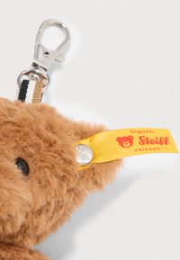 Tierno oso de peluche color marrón con un brillante botón plateado y una etiqueta amarilla que dice "Diseñado por Steiff Friends". Sujetado con un mosquetón de metal.