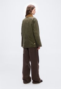 Γυναίκα που στέκεται πλάγια φορώντας μια χακί πράσινη ζακέτα με καφέ γιακά και φαρδιά, σκούρα καφέ παντελόνια, μπροστά σε απλό λευκό φόντο.