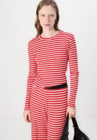 Czerwono-biały paskowany longsleeve z okrągłym dekoltem, zestawiony z pasującymi spodniami z czarnym paskiem, trzymany przez rękę.