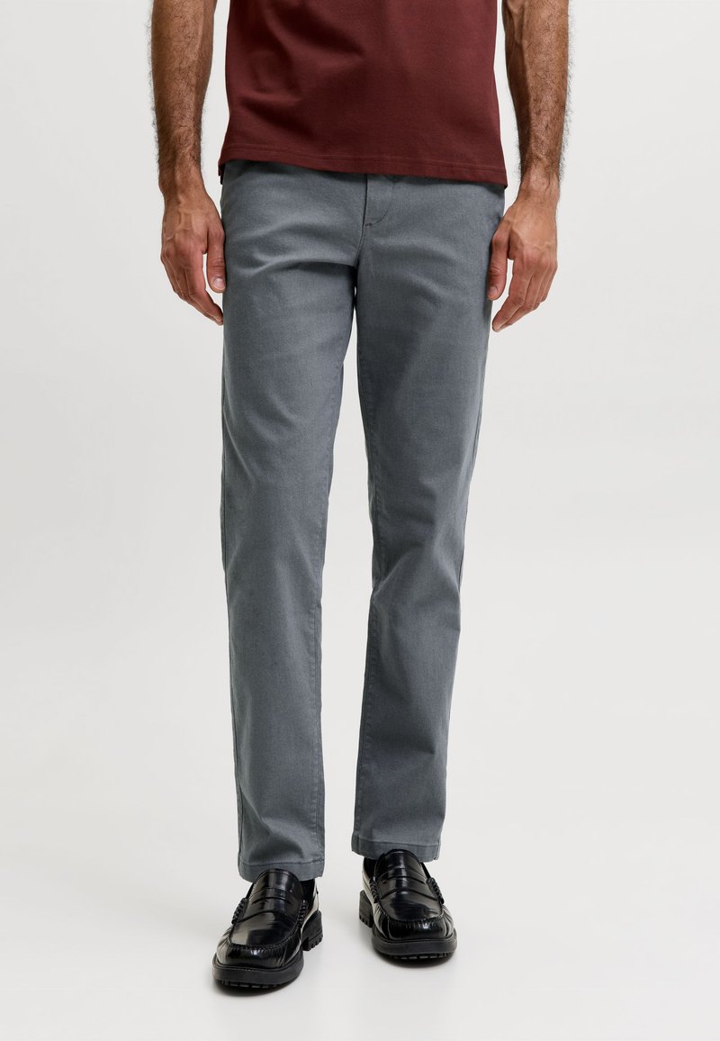 Homme portant un pantalon gris à jambes droites, des mocassins noirs et une chemise bordeaux, debout devant un fond clair uni.