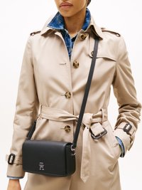 Schwarze, strukturierte Leder-Crossbody-Tasche mit silbernem Logo, aufgehängt an einem dünnen Riemen, getragen über einem beigen Trenchcoat mit Denim-Krag.
