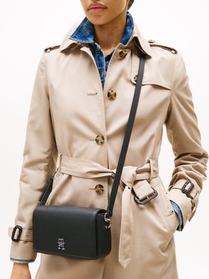 Zwarte gestructureerde leren crossbody tas met een zilveren logo, hangend aan een dunne strap, gedragen over een beige trenchcoat met een denim kraag.